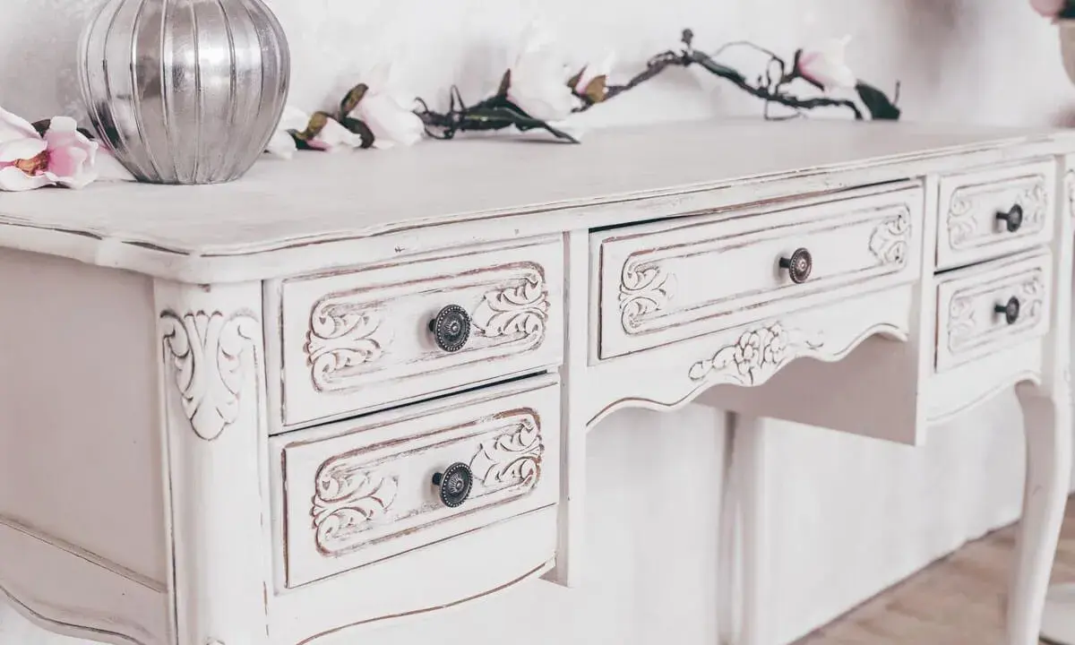 Toeletta in legno con **tecnica shabby a due colori**, decorata con intagli floreali e pomelli antichi. Un vaso d'argento e fiori rosa completano la scena.