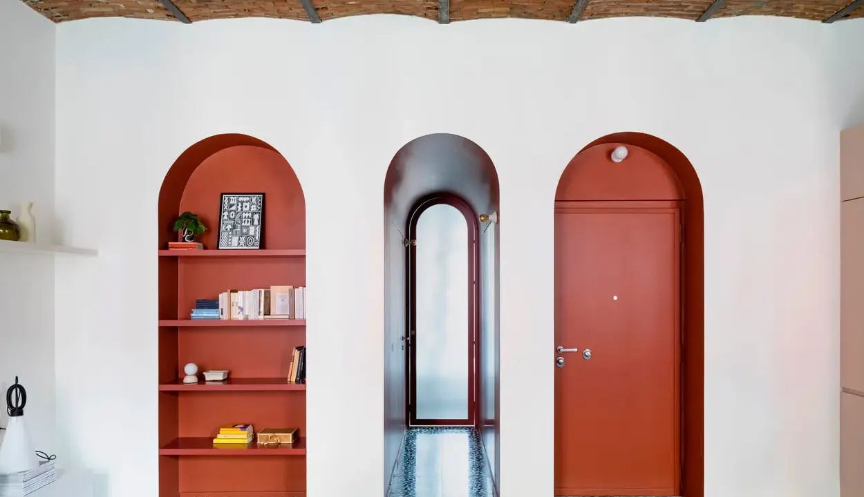 Una nicchia nel muro con mensole color terracotta ospita libri e decorazioni, accanto a un corridoio e una porta rossa.