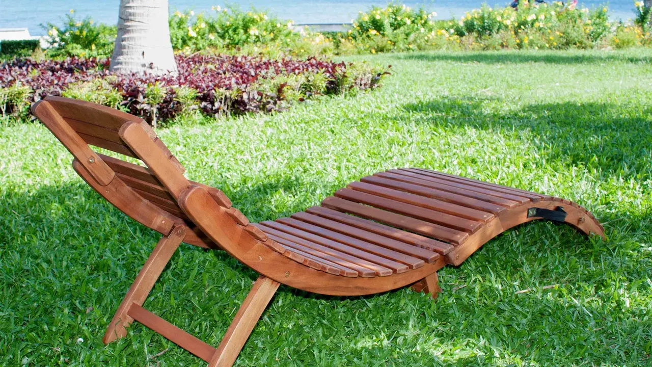 Chaise longue pieghevole in legno su prato verde, con mare e palme sullo sfondo. Ideale per rilassarsi all'aperto.