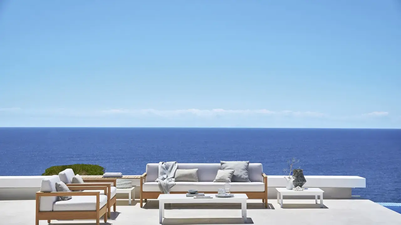 Un elegante divano Bali in legno e cuscini chiari, con vista sul mare blu intenso sotto un cielo limpido.
