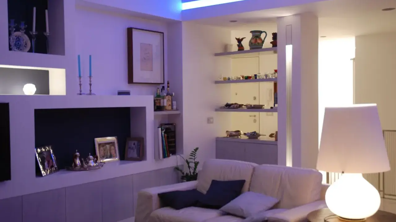 Soggiorno moderno con illuminazione soffusa, ideale per come illuminare un soggiorno con poca luce. Divano bianco, mensole con oggetti e lampada da tavolo.