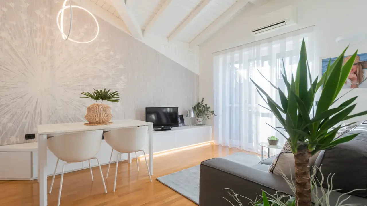 Luminoso appartamento mansardato con travi a vista, zona giorno con tavolo, sedie, TV e divano. Parete con carta da parati decorativa.
