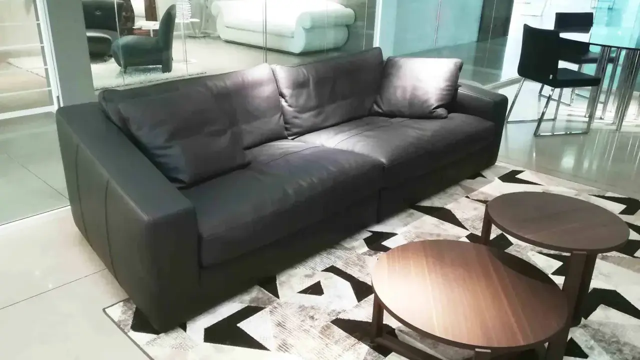 Un elegante divano angolare in pelle nera, con tavolini rotondi in legno, su un tappeto geometrico. Un frighetto è visibile in lontananza.