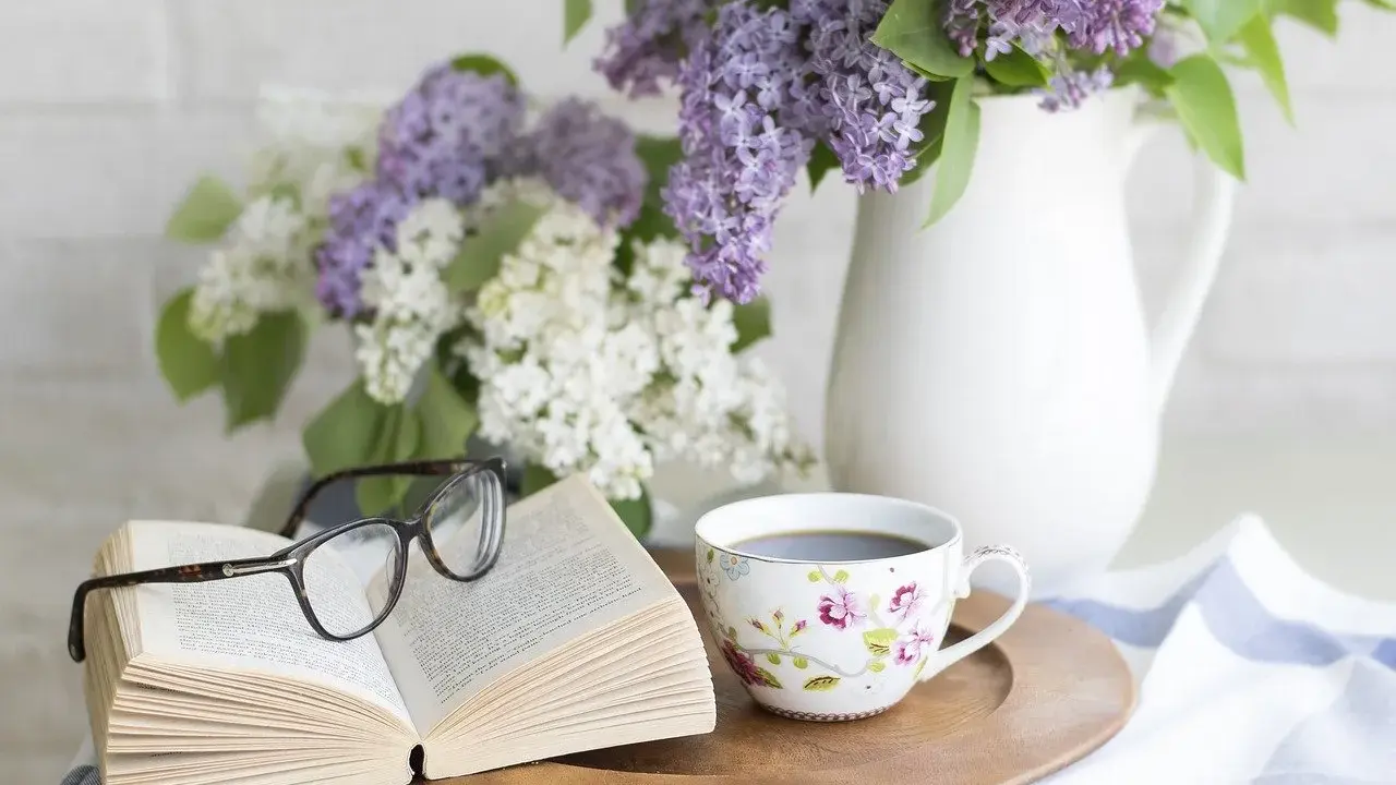 Un libro aperto, occhiali, tazza di caffè e fiori di lillà. Un'atmosfera rilassante, perfetta per pensare a come eliminare gli odori in casa.