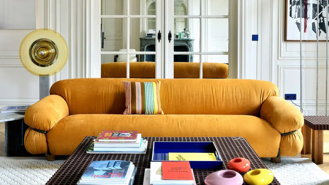 Divano senape con cuscino a righe colorate. Tavolino con libri e oggetti decorativi. Abbinamenti moderni e audaci.