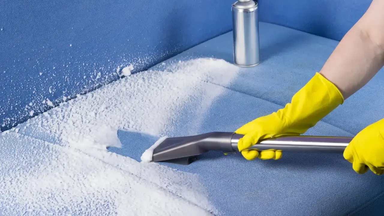 Mani con guanti gialli usano un aspiratore per pulire un divano in microfibra con schiuma detergente.