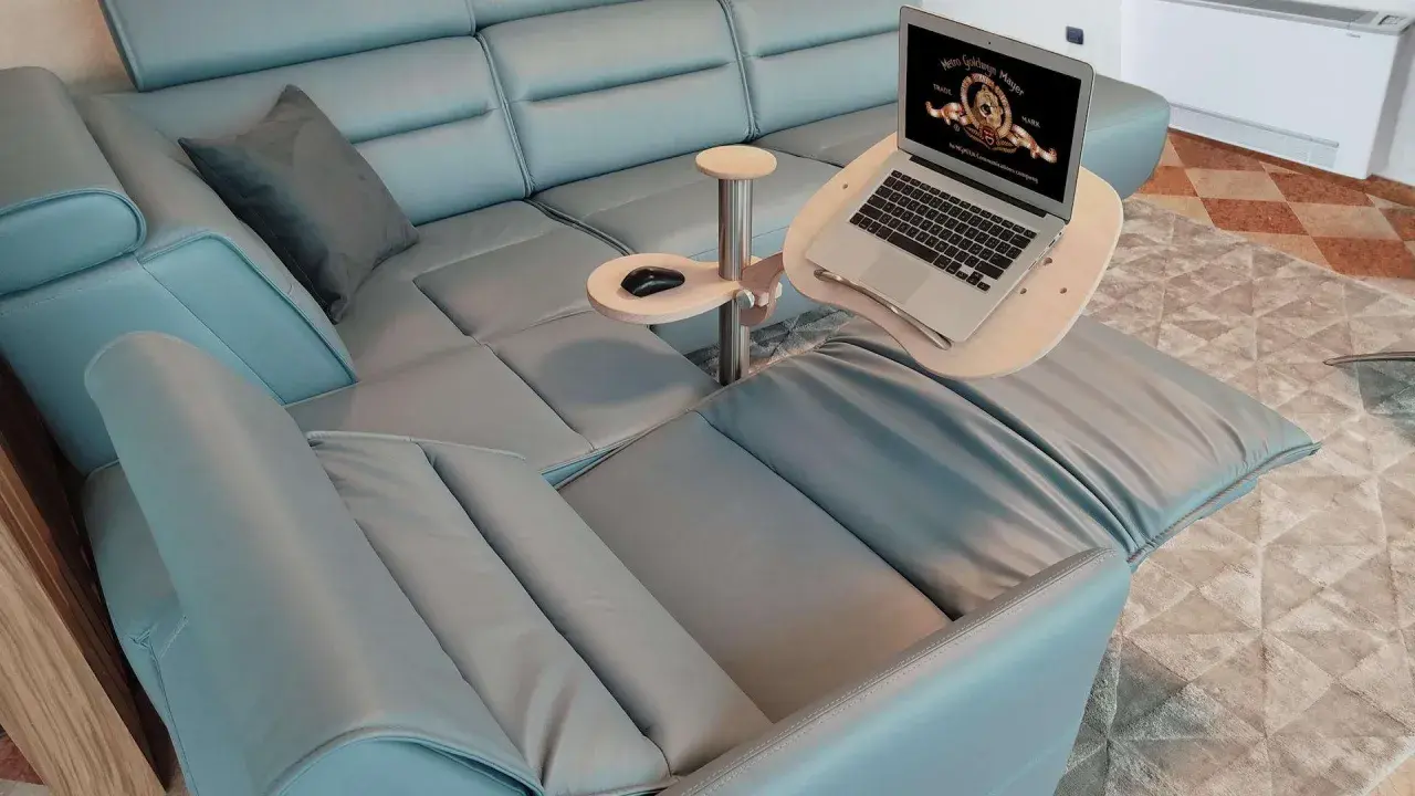 Comodo tavolino porta pc in legno e metallo, agganciato a un divano in pelle azzurra, con laptop aperto e mouse.
