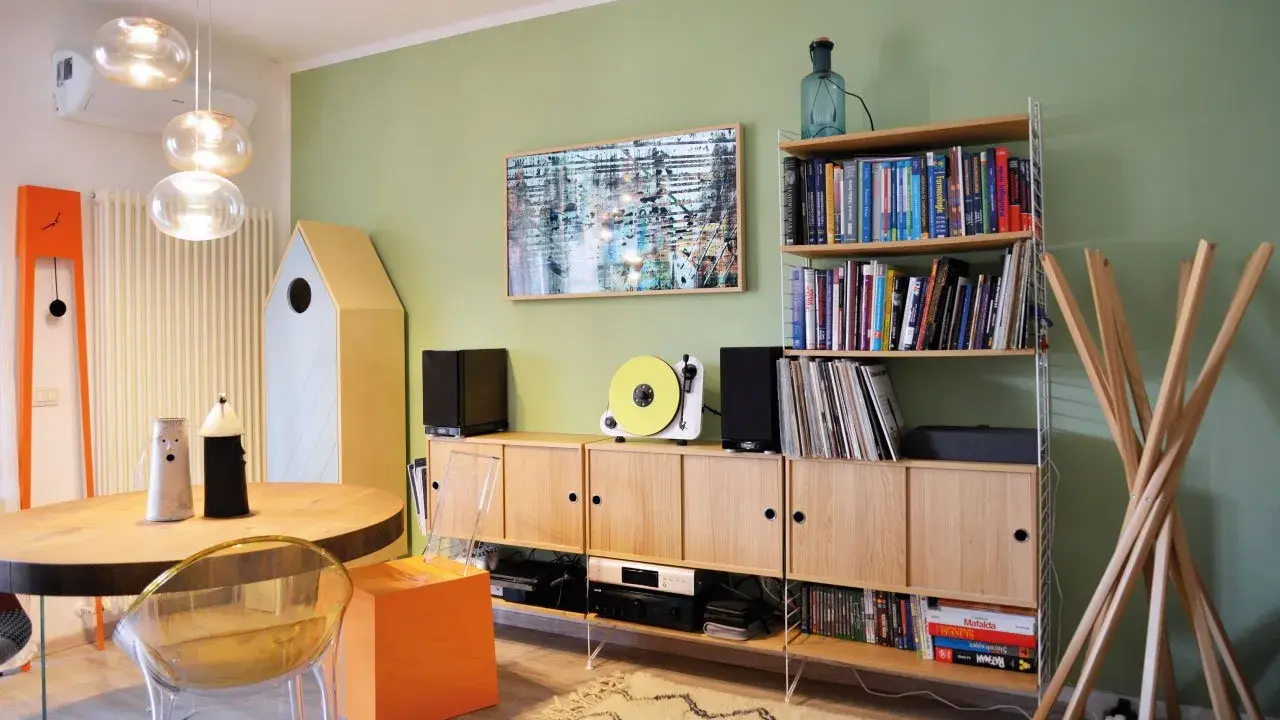 Soggiorno con pareti verde salvia, mobili in legno chiaro, giradischi giallo e un quadro astratto. I colori per pareti soggiorno sono rilassanti.