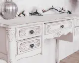 Toeletta in legno con **tecnica shabby a due colori**, decorata con intagli floreali e pomelli antichi. Un vaso d'argento e fiori rosa completano la scena.