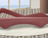 A pink velvet sofa with a unique curved design sits on a cream rug in a room with a geometric tiled floor and a view of the ocean.