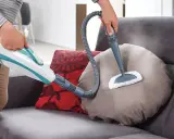 Persona pulisce un divano non sfoderabile con vaporetto, igienizzando i cuscini con vapore.