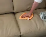 Mano che pulisce un divano in tessuto non sfoderabile con un panno arancione. Accanto, un panno grigio piegato.