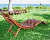 Chaise longue pieghevole in legno su prato verde, con mare e palme sullo sfondo. Ideale per rilassarsi all'aperto.