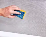 Mano che pulisce una zanzariera con una spugna blu e gialla. Un modo semplice per imparare come pulire le zanzariere.