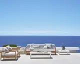 Un elegante divano Bali in legno e cuscini chiari, con vista sul mare blu intenso sotto un cielo limpido.