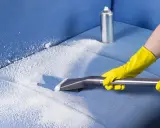 Mani con guanti gialli eseguono il lavaggio divano a secco con schiuma e aspiratore su tessuto blu.