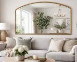 Ampio specchio dietro il divano, con cornice dorata, riflette una pianta, un lampadario e quadri.