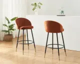 Due poltrone sgabello color ruggine, con gambe nere e dettagli dorati, in un ambiente moderno con piante e una barra.