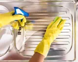 Mani con guanti gialli puliscono un lavello in acciaio con uno spray e una spugna, per far brillare l'acciaio.