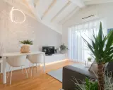 Luminoso appartamento mansardato con travi a vista, zona giorno con tavolo, sedie, TV e divano. Parete con carta da parati decorativa.