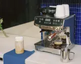 Un angolo caffè casa con macchina espresso, macinacaffè, tazzine e un bicchiere di cappuccino.