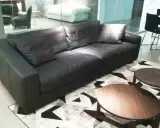 Un elegante divano angolare in pelle nera, con tavolini rotondi in legno, su un tappeto geometrico. Un frighetto è visibile in lontananza.