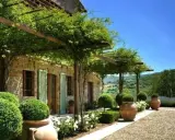Splendide idee case di campagna esterni: un pergolato verde ombreggia una casa in pietra con persiane verdi, circondata da piante in vaso e un paesaggio collinare.