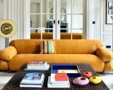 Divano senape con cuscino a righe colorate. Tavolino con libri e oggetti decorativi. Abbinamenti moderni e audaci.