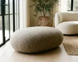 Un morbido pouf dal design organico, rivestito in tessuto grezzo, si abbina a un divano bouclé e a un vaso in terracotta.