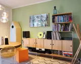 Soggiorno con pareti verde salvia, mobili in legno chiaro, giradischi giallo e un quadro astratto. I colori per pareti soggiorno sono rilassanti.