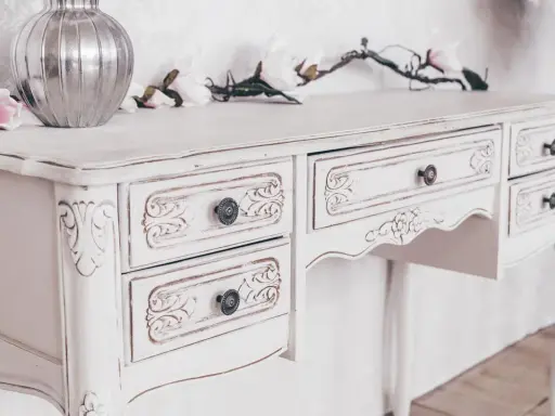 Toeletta in legno con **tecnica shabby a due colori**, decorata con intagli floreali e pomelli antichi. Un vaso d'argento e fiori rosa completano la scena.
