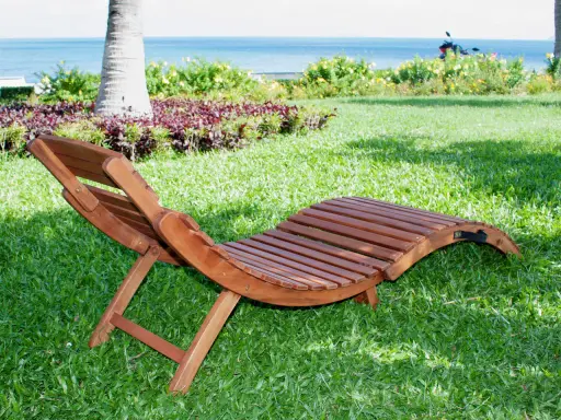 Chaise longue pieghevole in legno su prato verde, con mare e palme sullo sfondo. Ideale per rilassarsi all'aperto.