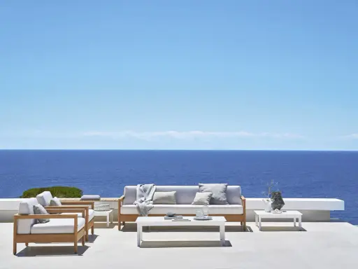 Un elegante divano Bali in legno e cuscini chiari, con vista sul mare blu intenso sotto un cielo limpido.