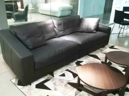 Un elegante divano angolare in pelle nera, con tavolini rotondi in legno, su un tappeto geometrico. Un frighetto è visibile in lontananza.