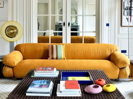 Divano senape con cuscino a righe colorate. Tavolino con libri e oggetti decorativi. Abbinamenti moderni e audaci.