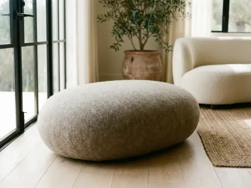 Un morbido pouf dal design organico, rivestito in tessuto grezzo, si abbina a un divano bouclé e a un vaso in terracotta.
