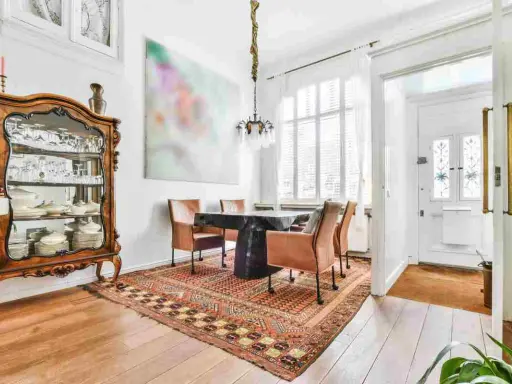 Soggiorno con arredamento misto moderno e antico: vetrina in legno intarsiato, tavolo scuro con sedie in pelle, tappeto persiano e quadro astratto.