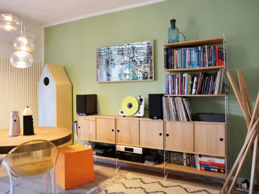 Soggiorno con pareti verde salvia, mobili in legno chiaro, giradischi giallo e un quadro astratto. I colori per pareti soggiorno sono rilassanti.