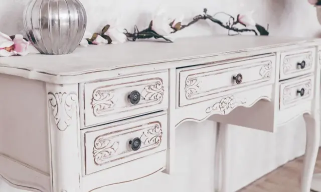 Toeletta in legno con **tecnica shabby a due colori**, decorata con intagli floreali e pomelli antichi. Un vaso d'argento e fiori rosa completano la scena.