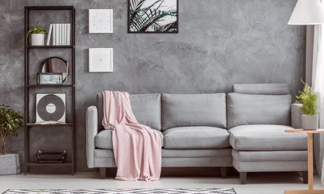 Un divano grigio con coperta rosa, una libreria nera con piante e vinile, e un tavolino di legno: ecco cosa mettere vicino al divano per un tocco di stile.