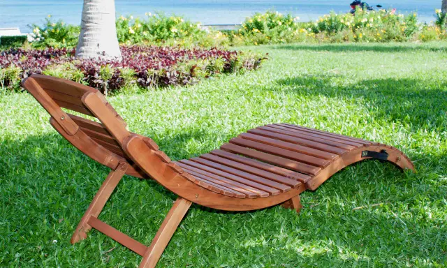 Chaise longue pieghevole in legno su prato verde, con mare e palme sullo sfondo. Ideale per rilassarsi all'aperto.