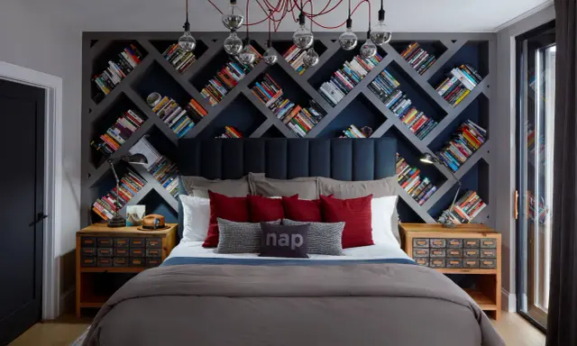 Camera da letto con un'imponente parete di librerie stravaganti a rombi, piene di libri colorati. Un letto accogliente con cuscini rossi e grigi.