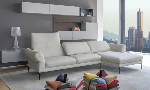 Un divano bianco in pelle, con cuscini colorati sparsi sul tappeto, davanti a una finestra con vista sulla città al tramonto. L'alba calia illumina la scena.