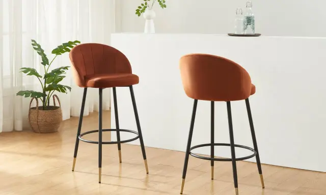 Due poltrone sgabello color ruggine, con gambe nere e dettagli dorati, in un ambiente moderno con piante e una barra.
