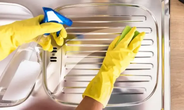 Mani con guanti gialli puliscono un lavello in acciaio con uno spray e una spugna, per far brillare l'acciaio.