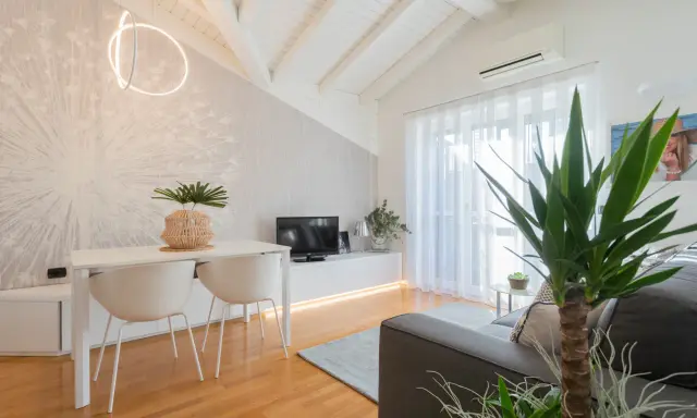 Luminoso appartamento mansardato con travi a vista, zona giorno con tavolo, sedie, TV e divano. Parete con carta da parati decorativa.