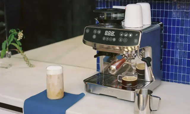 Un angolo caffè casa con macchina espresso, macinacaffè, tazzine e un bicchiere di cappuccino.
