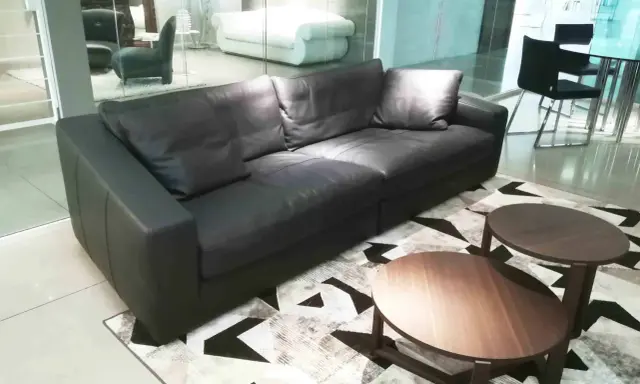 Un elegante divano angolare in pelle nera, con tavolini rotondi in legno, su un tappeto geometrico. Un frighetto è visibile in lontananza.
