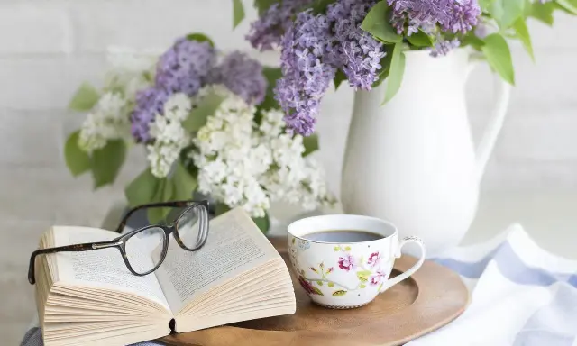 Un libro aperto, occhiali, tazza di caffè e fiori di lillà. Un'atmosfera rilassante, perfetta per pensare a come eliminare gli odori in casa.