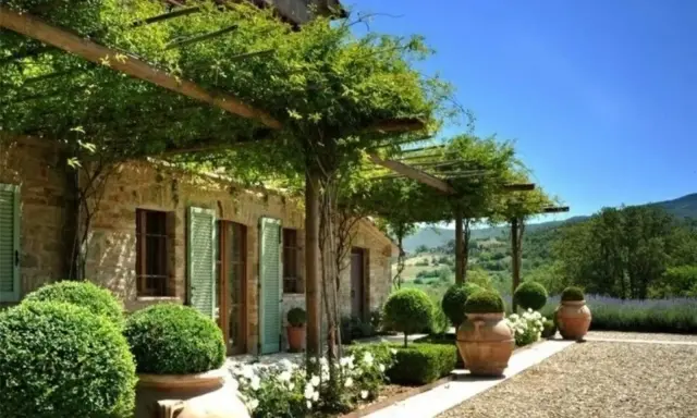 Splendide idee case di campagna esterni: un pergolato verde ombreggia una casa in pietra con persiane verdi, circondata da piante in vaso e un paesaggio collinare.
